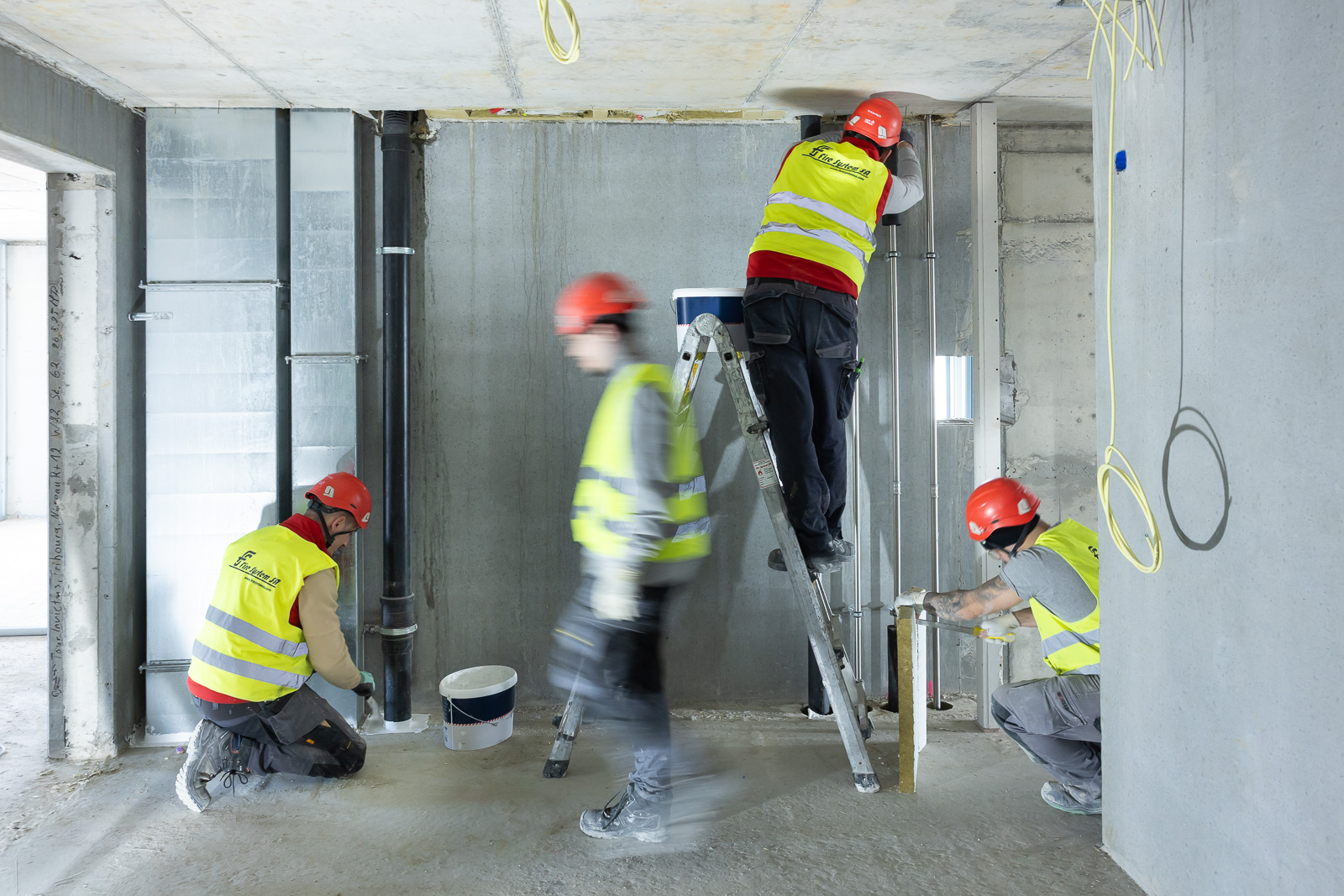 Fire System SA workers in hi-vis vests and red helmets install ductwork and conduits in a bare concrete interior under construction, one on a stepladder reaching to the ceiling while two others work at floor level.