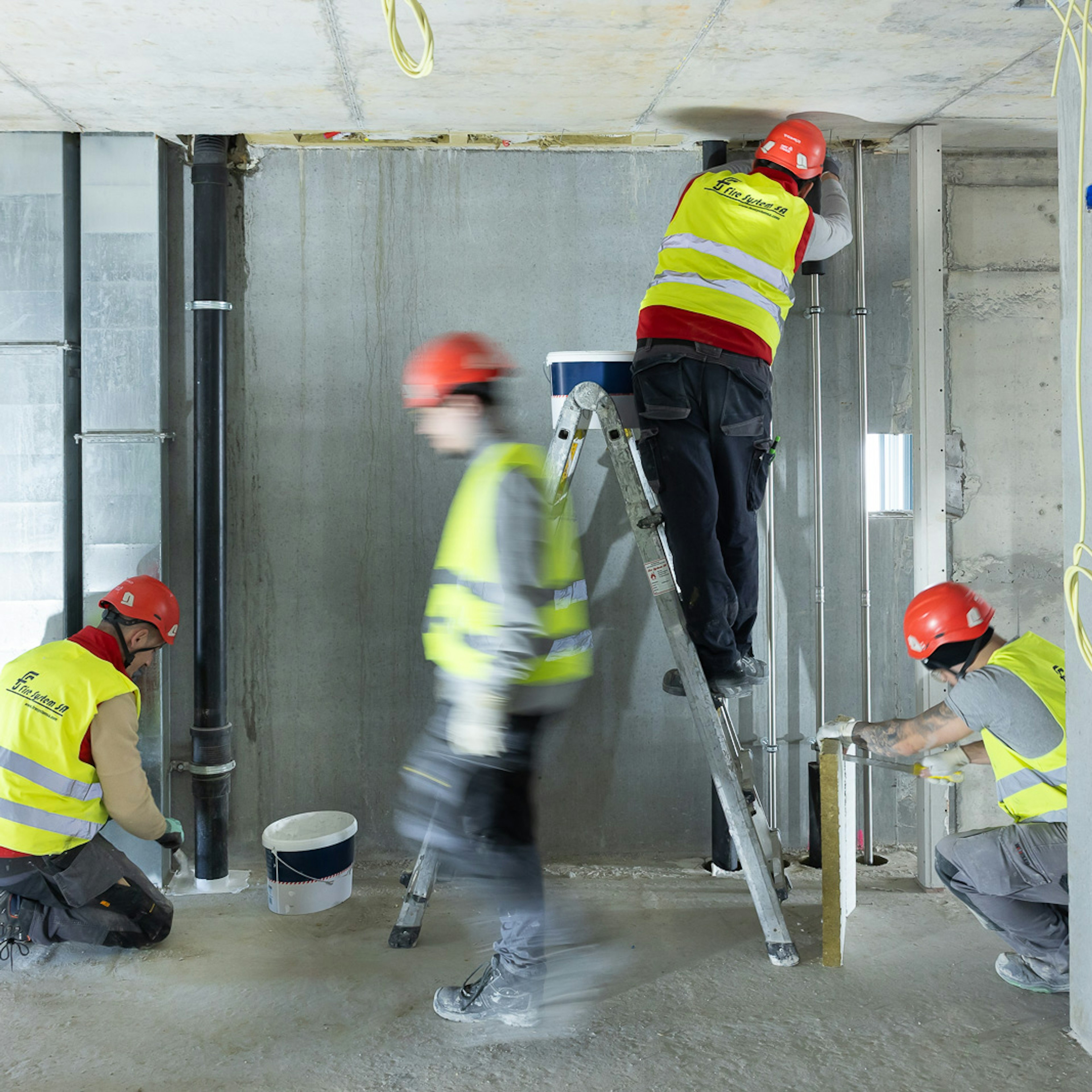 Des employés de Fire System SA, vêtus de gilets haute visibilité et de casques rouges, installent des conduits et des gaines dans un espace intérieur en béton brut en cours de construction ; l'un d'entre eux est perché sur un escabeau qui atteint le plafond, tandis que deux autres travaillent au niveau du sol.