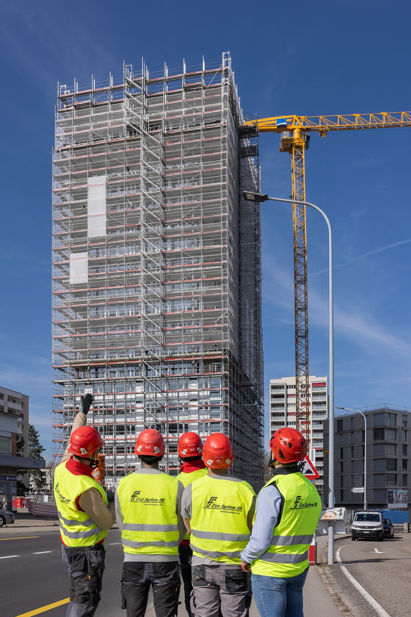 Five Fire System SA workers in hi-vis vests and red helmets stand with their backs to the camera, looking up at a scaffolded high-rise under construction, one pointing toward the top as a tower crane rises alongside.
