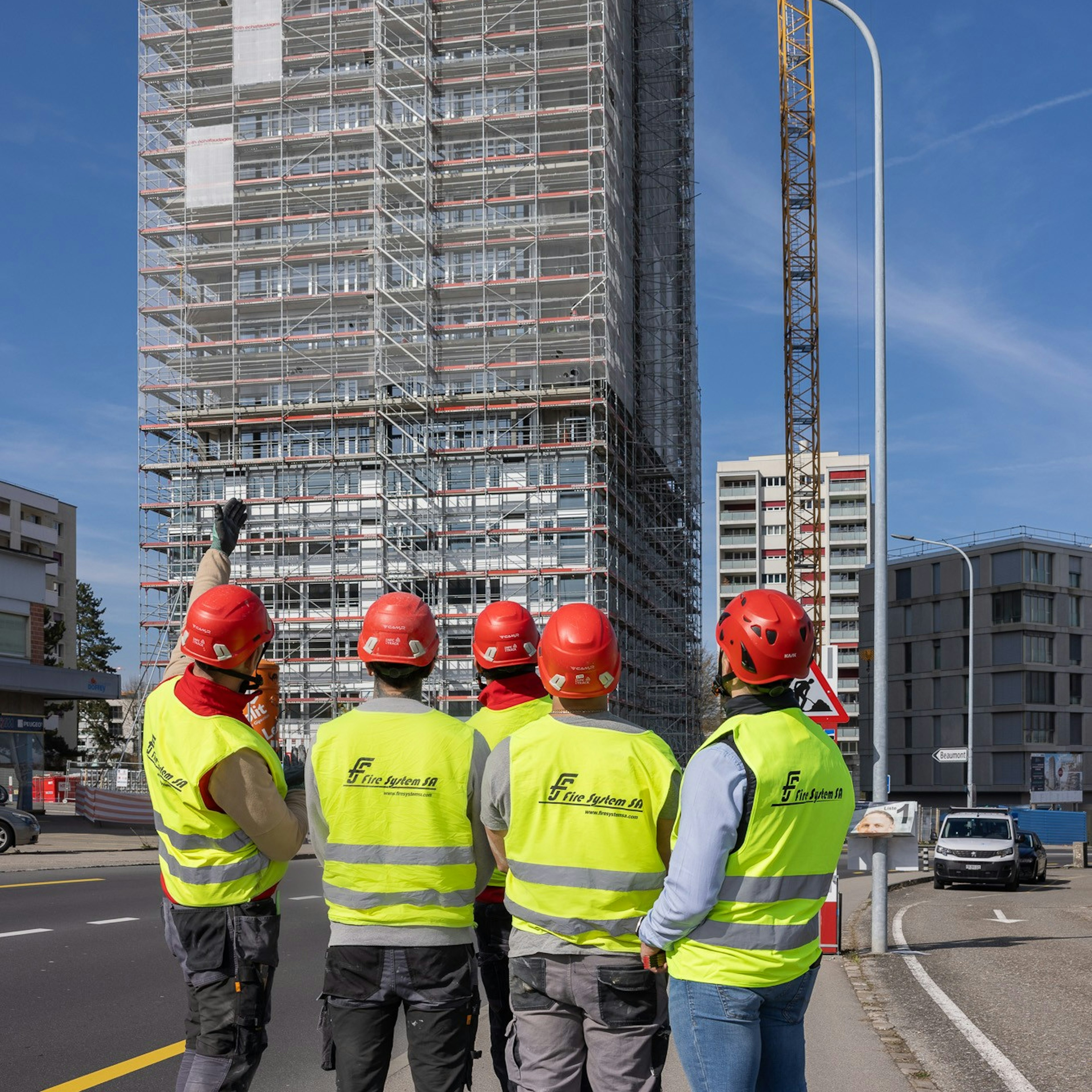 Cinq employés de Fire System SA, vêtus de gilets haute visibilité et de casques rouges, se tiennent dos à la caméra, le regard levé vers un gratte-ciel en chantier entouré d'échafaudages ; l'un d'eux pointe du doigt le sommet tandis qu'une grue à tour s'élève à côté.