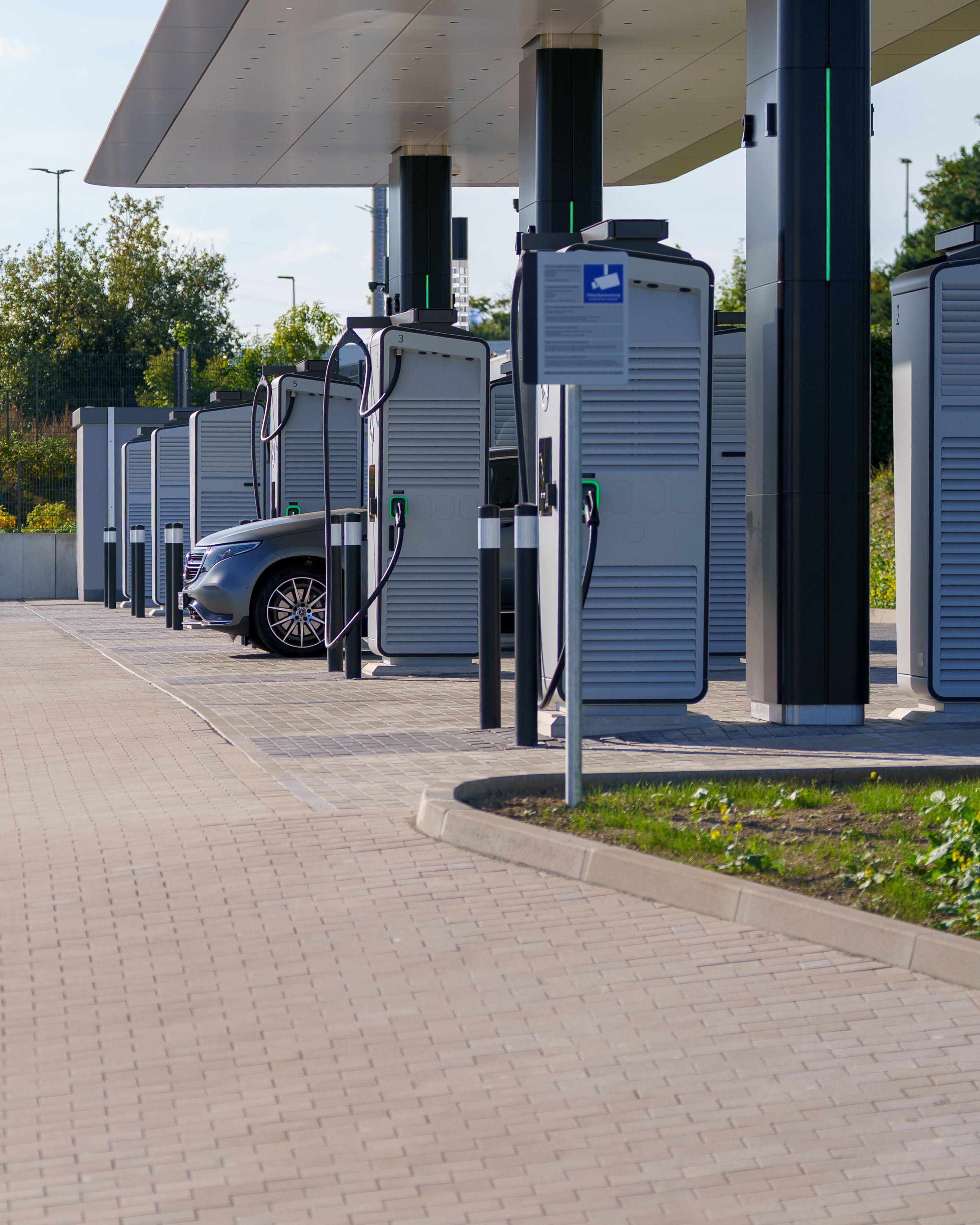 Une rangée de bornes de recharge rapide haute puissance pour véhicules électriques est installée sous un auvent sur un parvis pavé ; une voiture électrique argentée est branchée et en cours de recharge à l'une des bornes numérotées.