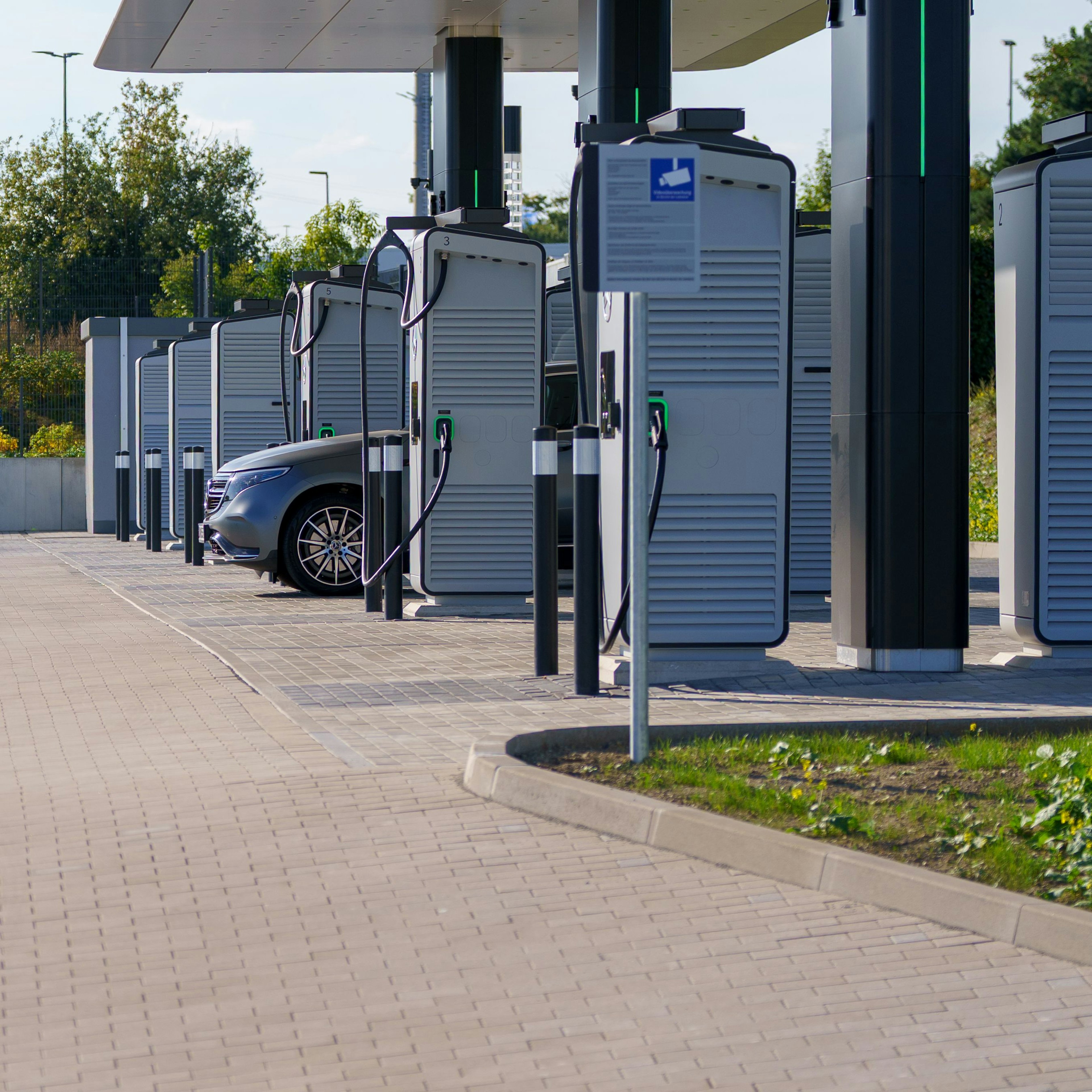 Une rangée de bornes de recharge rapide haute puissance pour véhicules électriques est installée sous un auvent sur un parvis pavé ; une voiture électrique argentée est branchée et en cours de recharge à l'une des bornes numérotées.