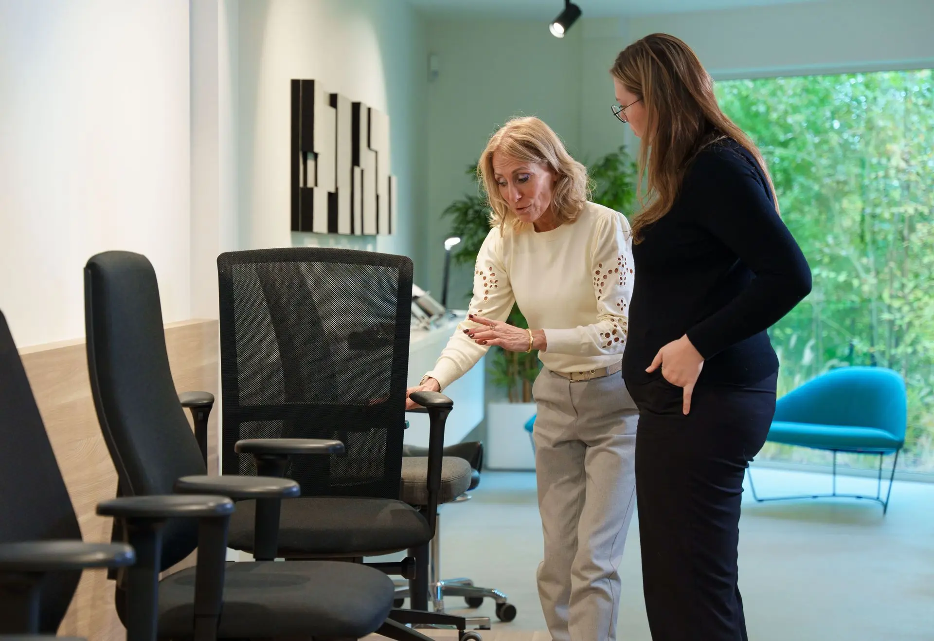 In a bright showroom, one person points out the features of a black mesh-back ergonomic office chair to another, with more chairs alongside.