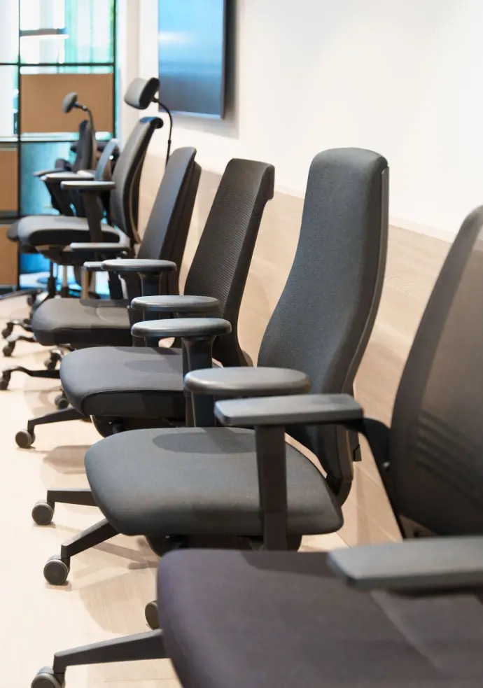 A row of dark grey ergonomic office chairs with adjustable armrests lined up along a light wood-panelled meeting-room wall.