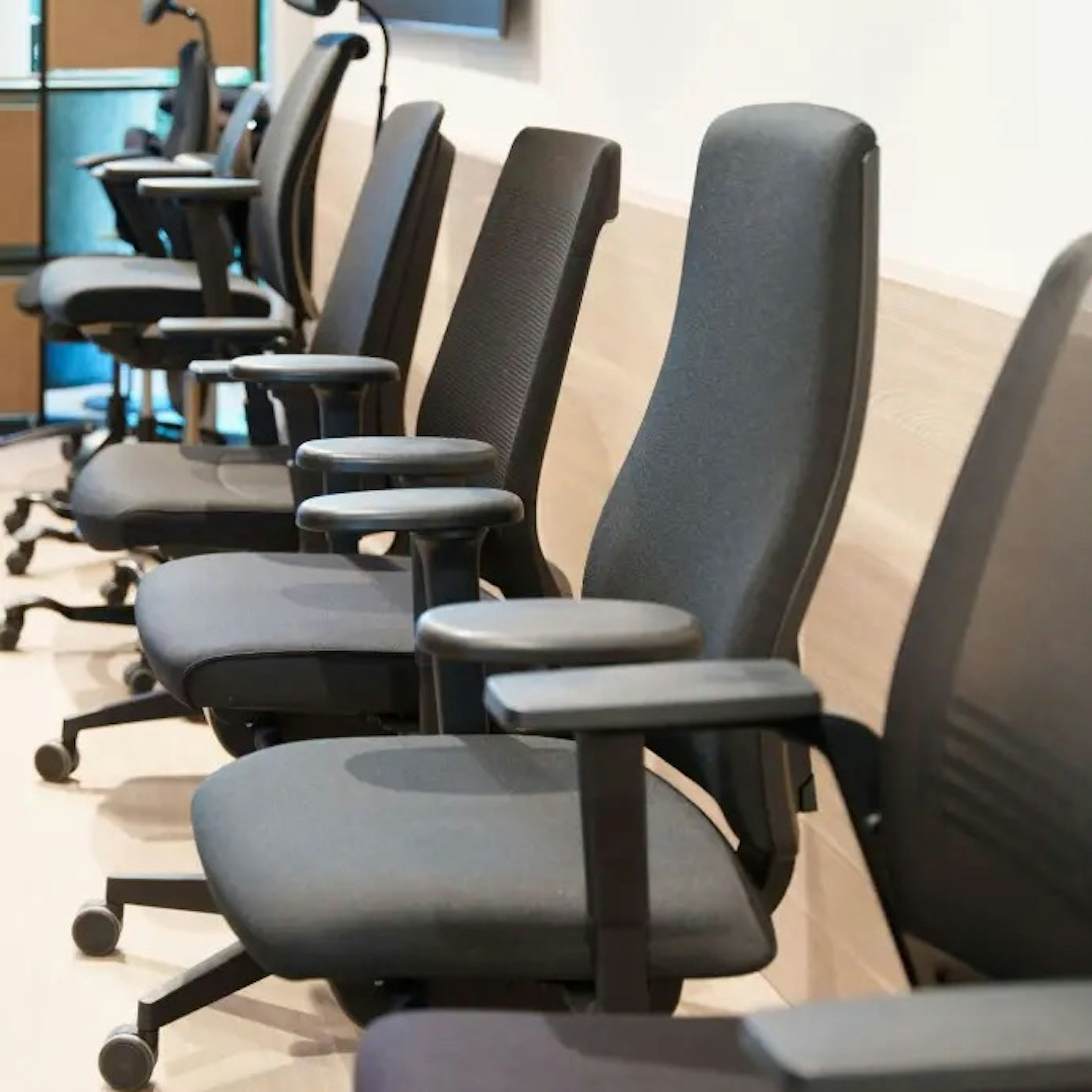 A row of dark grey ergonomic office chairs with adjustable armrests lined up along a light wood-panelled meeting-room wall.