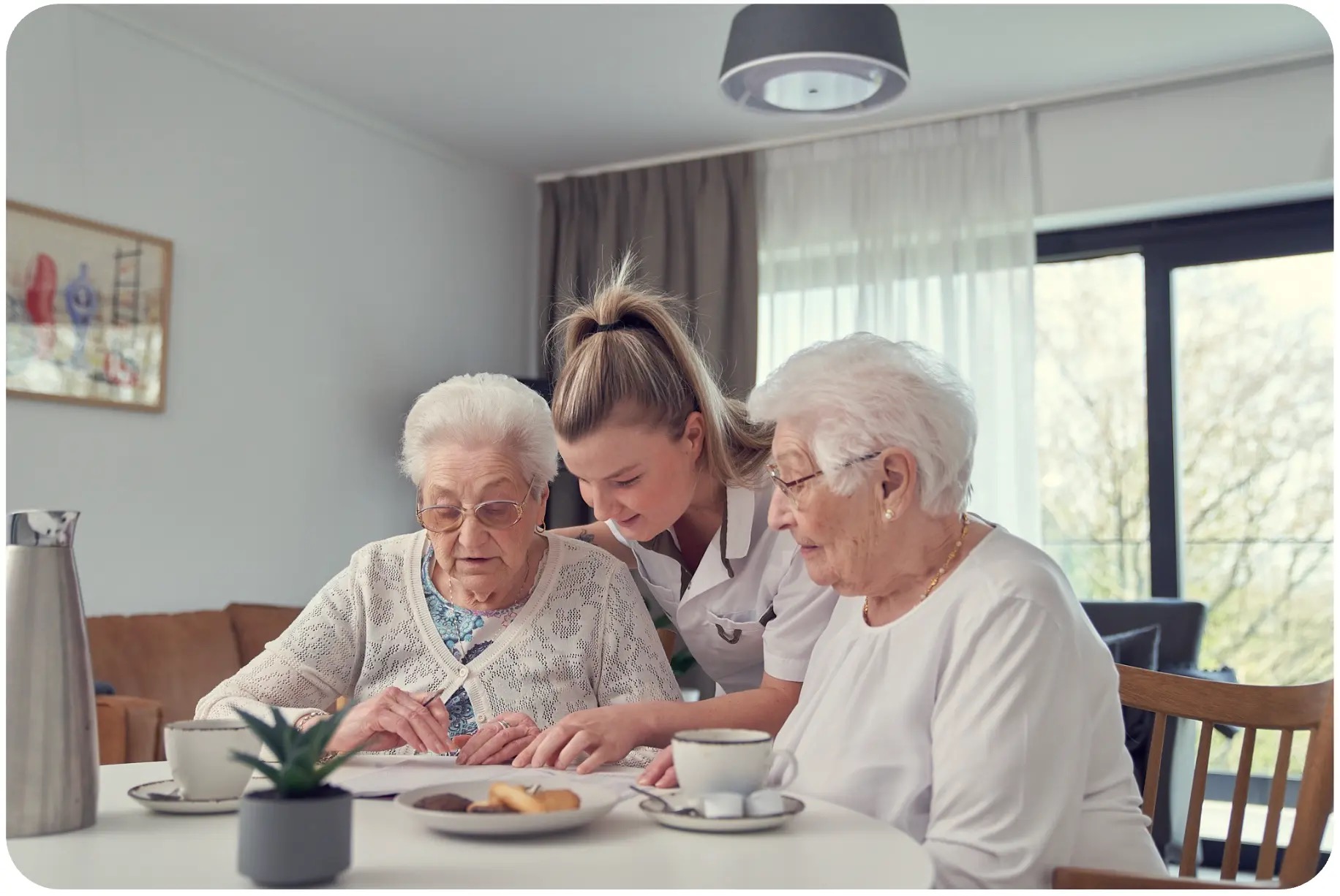 Een verzorger helpt twee bejaarde vrouwen aan een tafel, terwijl boven hen aan het plafond een Nobi-slimme lamp hangt.