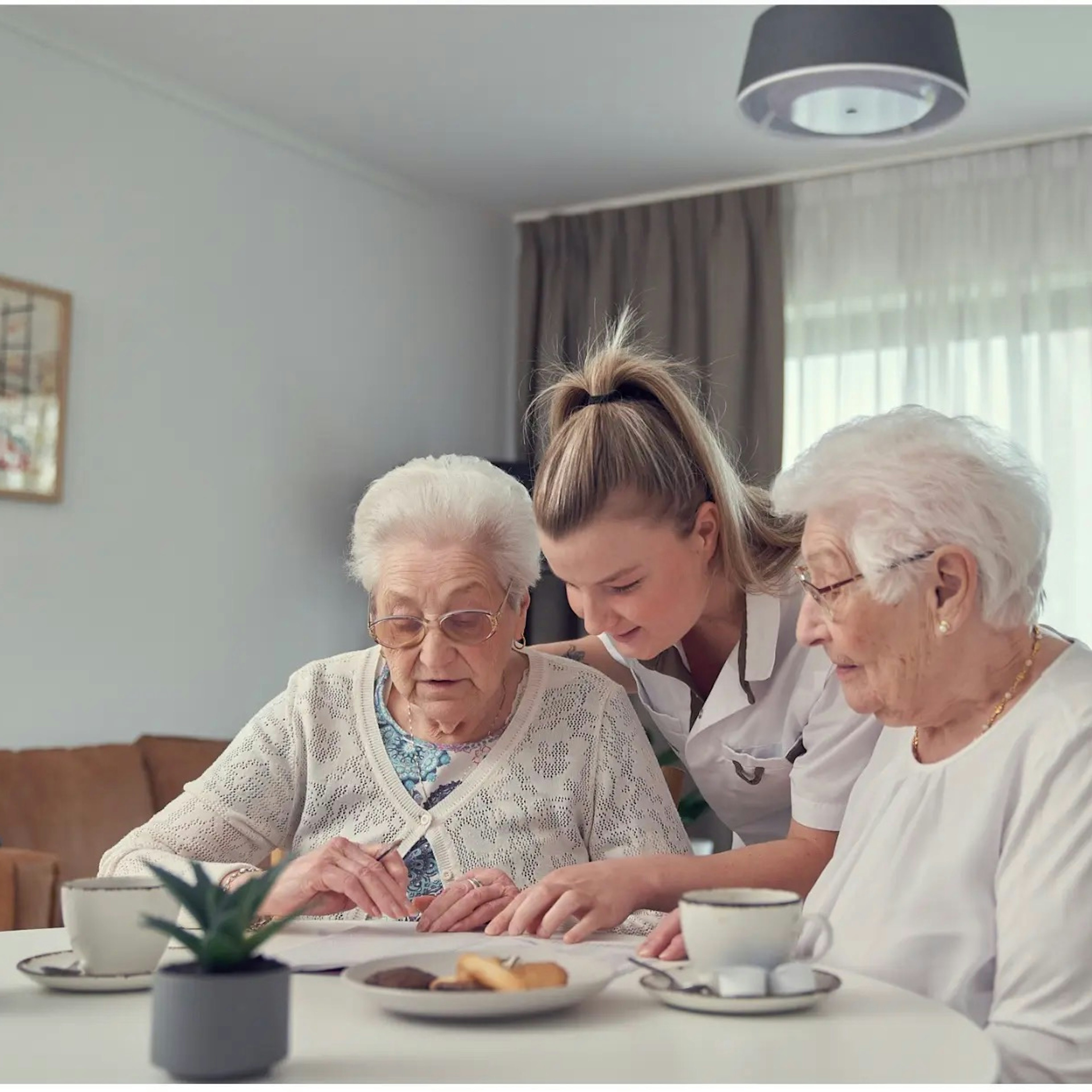 Een verzorger helpt twee bejaarde vrouwen aan een tafel, terwijl boven hen aan het plafond een Nobi-slimme lamp hangt.