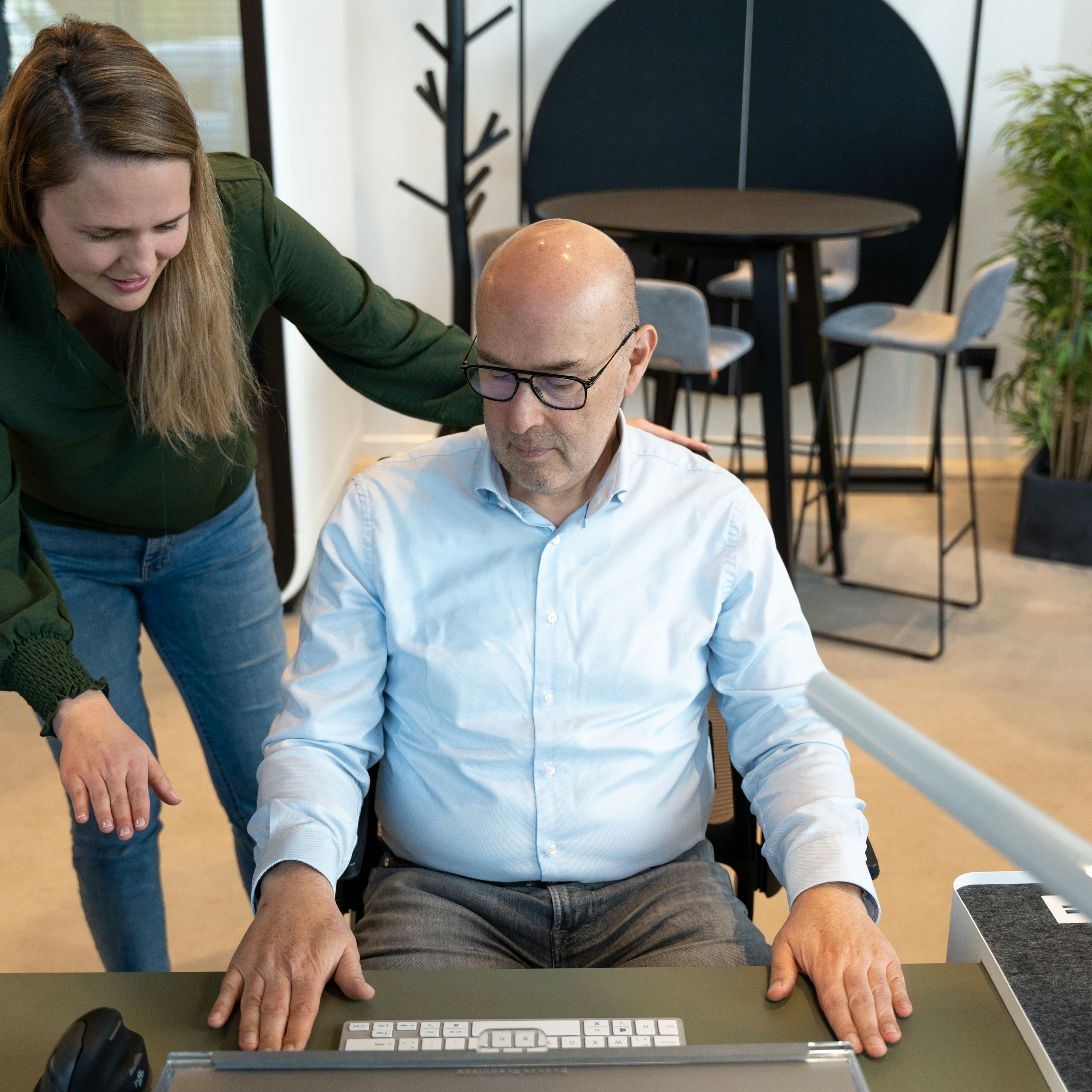Un collègue debout se penche pour aider un homme assis à un poste de travail ergonomique équipé d'un clavier divisé en deux parties et d'une souris verticale, dans un bureau lumineux agrémenté de plantes.