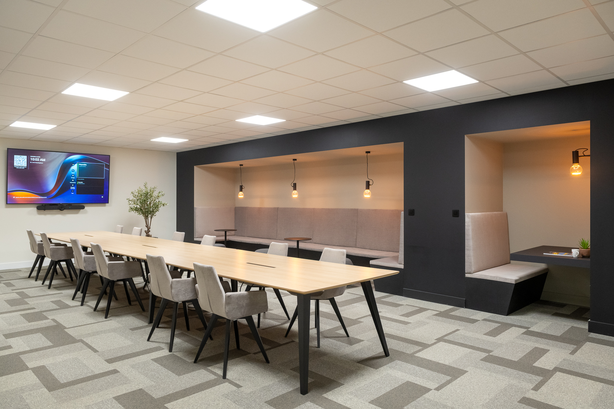 « Salle de réunion moderne équipée d'une longue table en bois clair et de chaises rembourrées grises, d'un écran mural et d'un coin salon rembourré adossé à un mur d'accent foncé. »