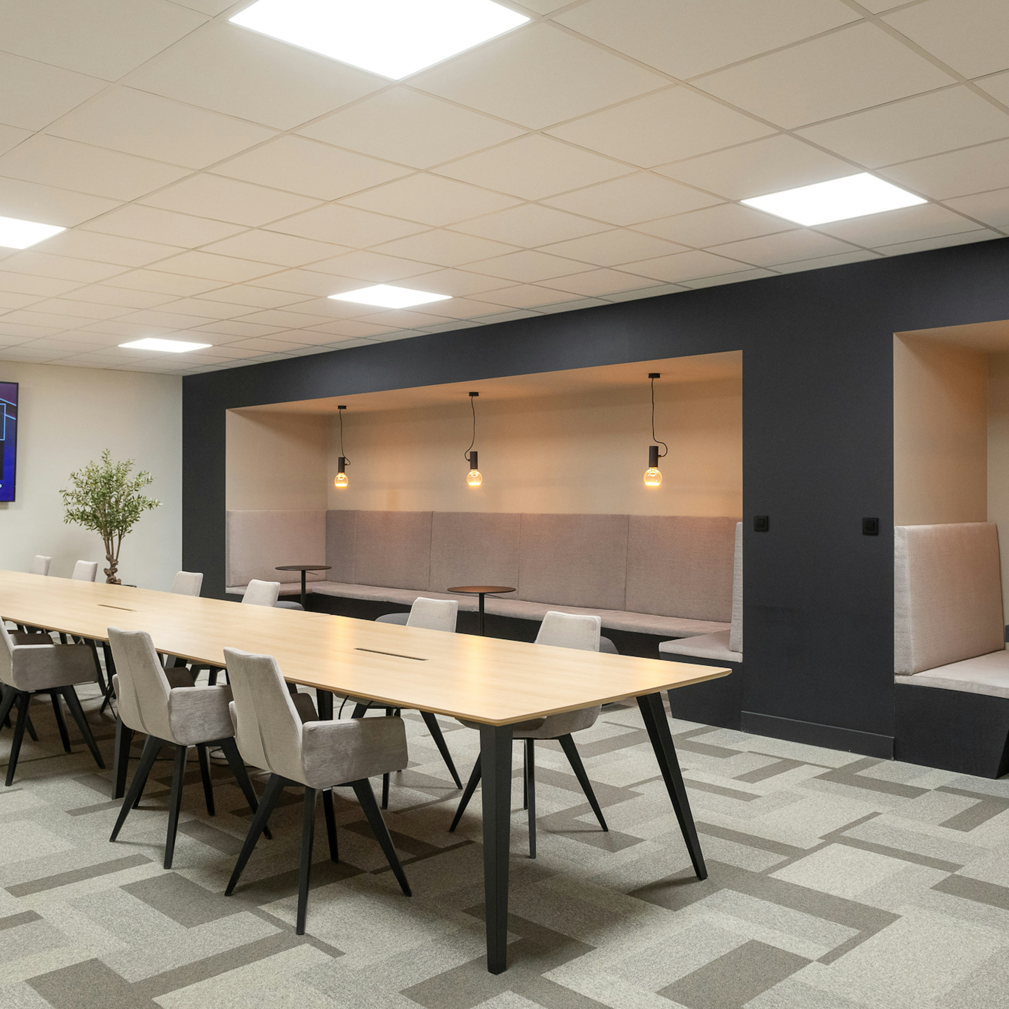 « Salle de réunion moderne équipée d'une longue table en bois clair et de chaises rembourrées grises, d'un écran mural et d'un coin salon rembourré adossé à un mur d'accent foncé. »