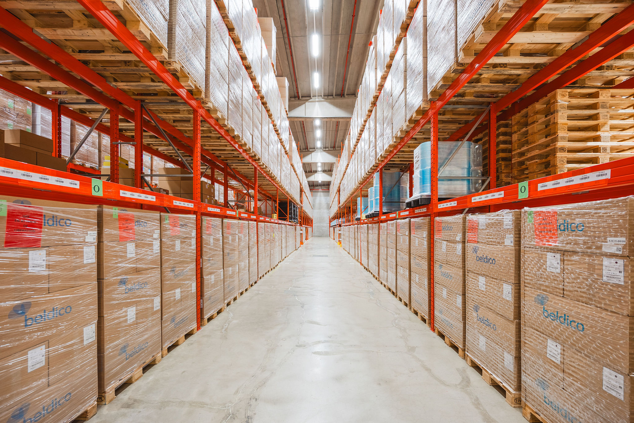 Een lange magazijngang met aan beide zijden hoge oranje palletstellingen, volledig gevuld met gewikkelde Beldico-dozen en pallets.