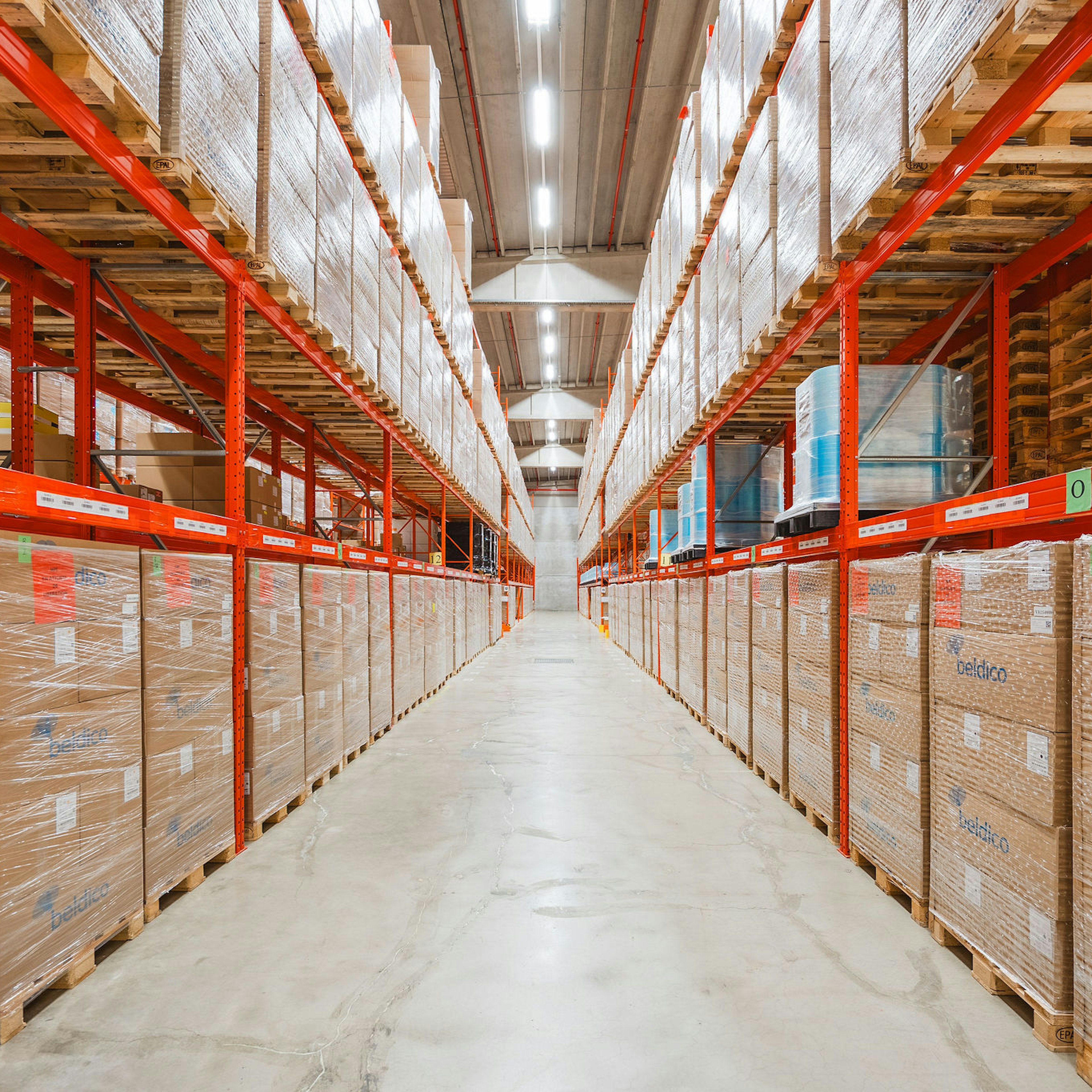 Een lange magazijngang met aan beide zijden hoge oranje palletstellingen, volledig gevuld met gewikkelde Beldico-dozen en pallets.