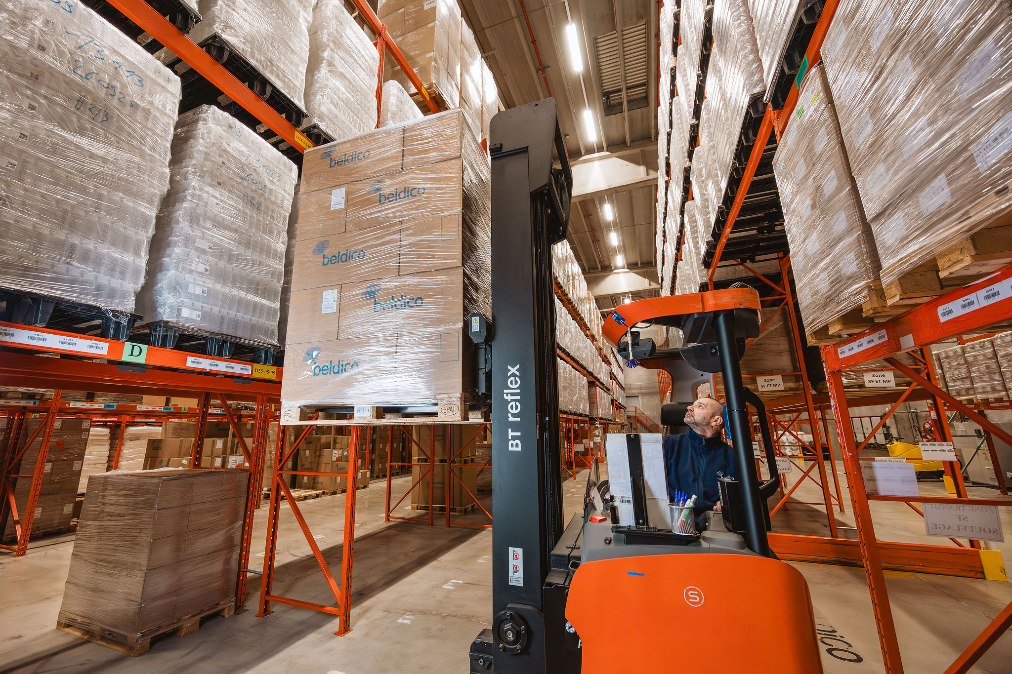 Een magazijnmedewerker bestuurt een oranje BT Reflex reachtruck en tilt een gewikkelde pallet met Beldico-dozen in hoge stellingen.