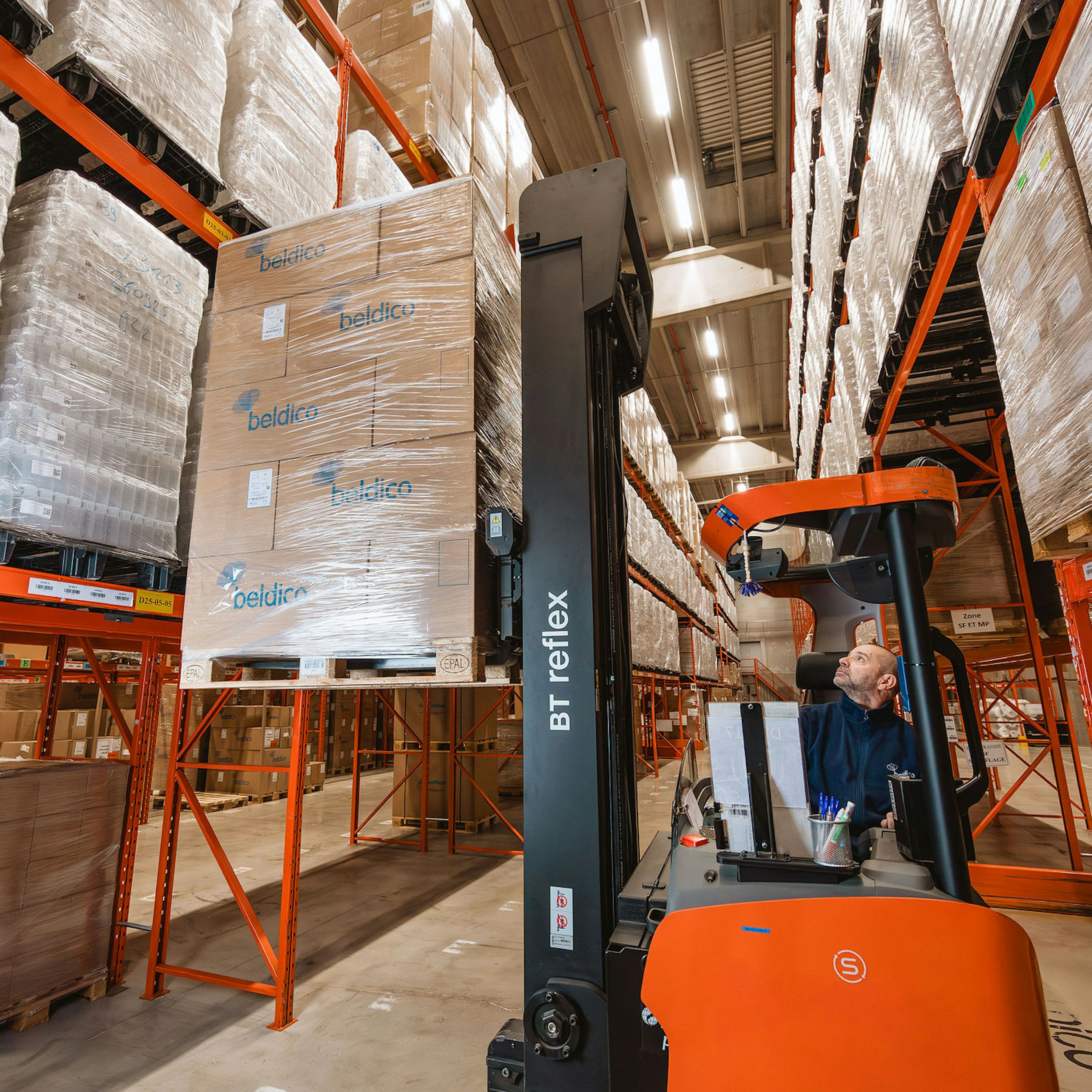 Een magazijnmedewerker bestuurt een oranje BT Reflex reachtruck en tilt een gewikkelde pallet met Beldico-dozen in hoge stellingen.