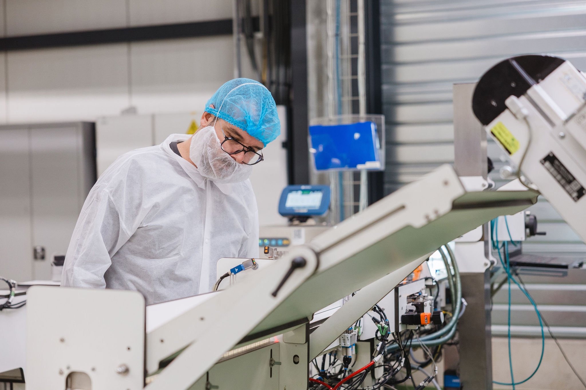 Een medewerker in een witte overall, haarnetje, mondmasker en bril bedient een geautomatiseerde verpakkingslijn in een gecontroleerde productieomgeving.