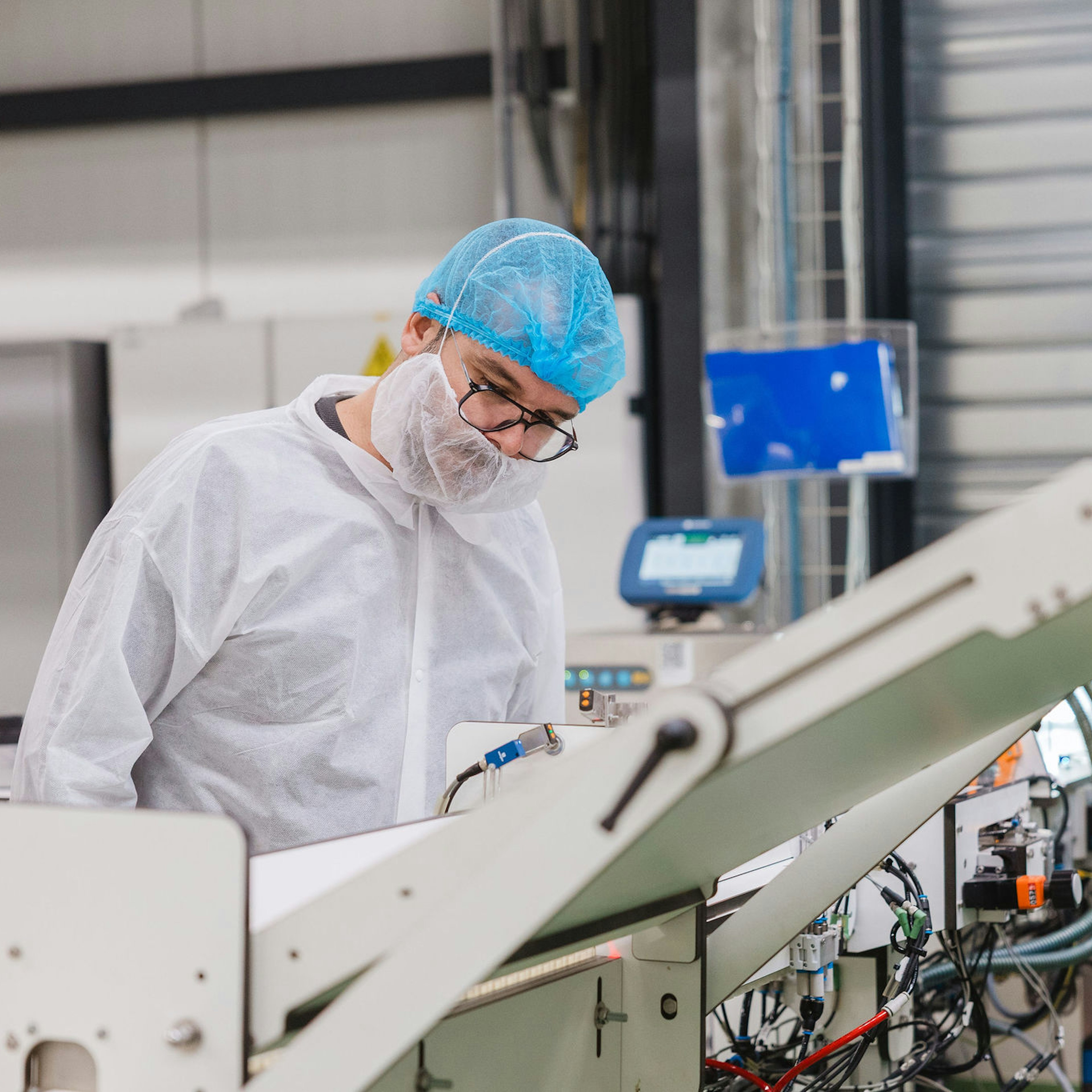 Een medewerker in een witte overall, haarnetje, mondmasker en bril bedient een geautomatiseerde verpakkingslijn in een gecontroleerde productieomgeving.