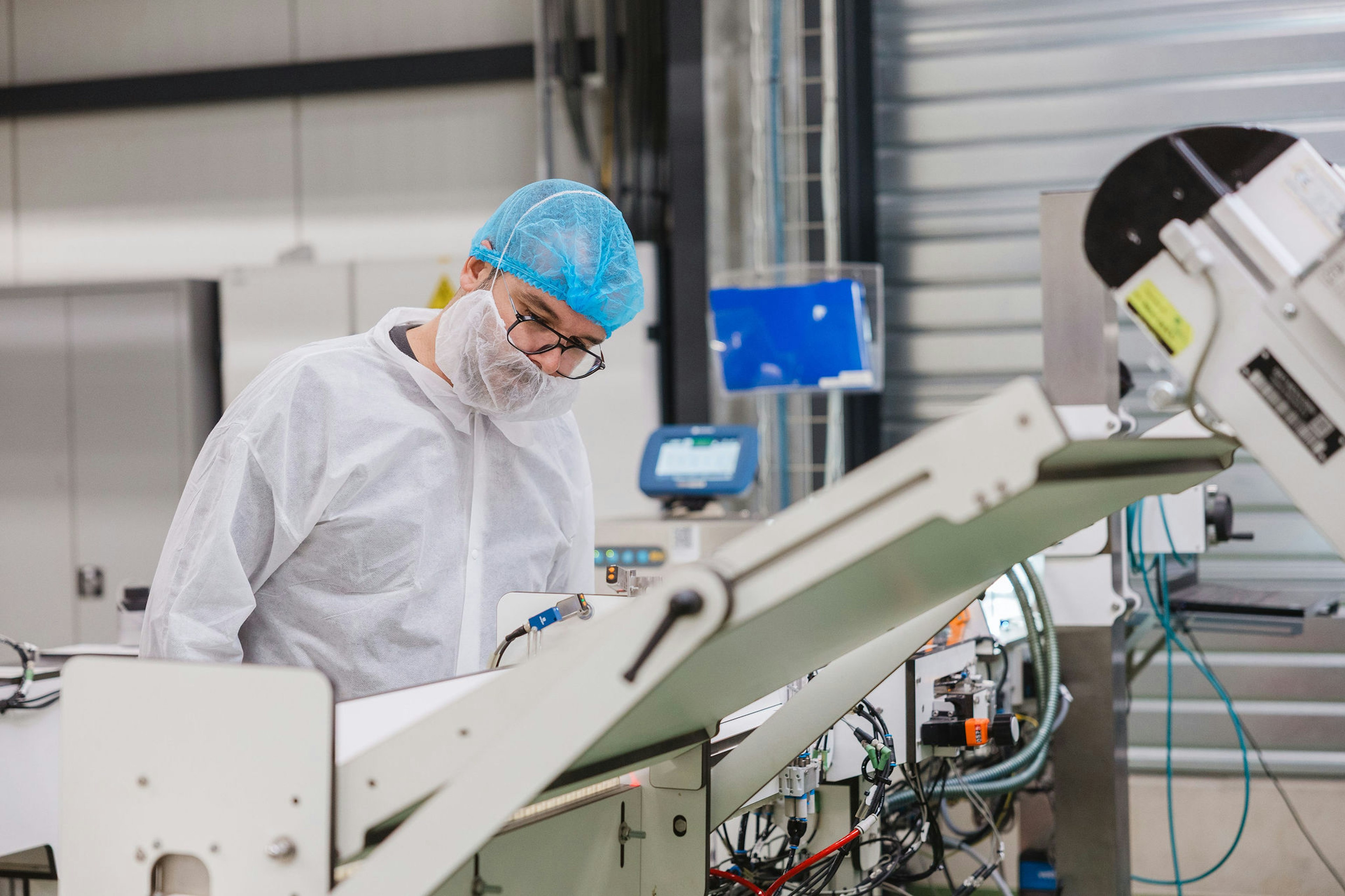 Een medewerker in een witte overall, haarnetje, mondmasker en bril bedient een geautomatiseerde verpakkingslijn in een gecontroleerde productieomgeving.