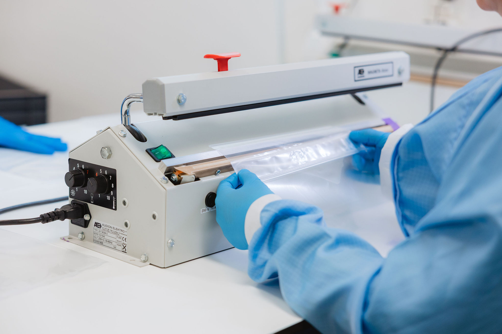 Een medewerker in een blauwe beschermende jas en nitrilhandschoenen sealt een transparant plastic zakje met een Audion Elektro impulssealer in een schone productieomgeving.