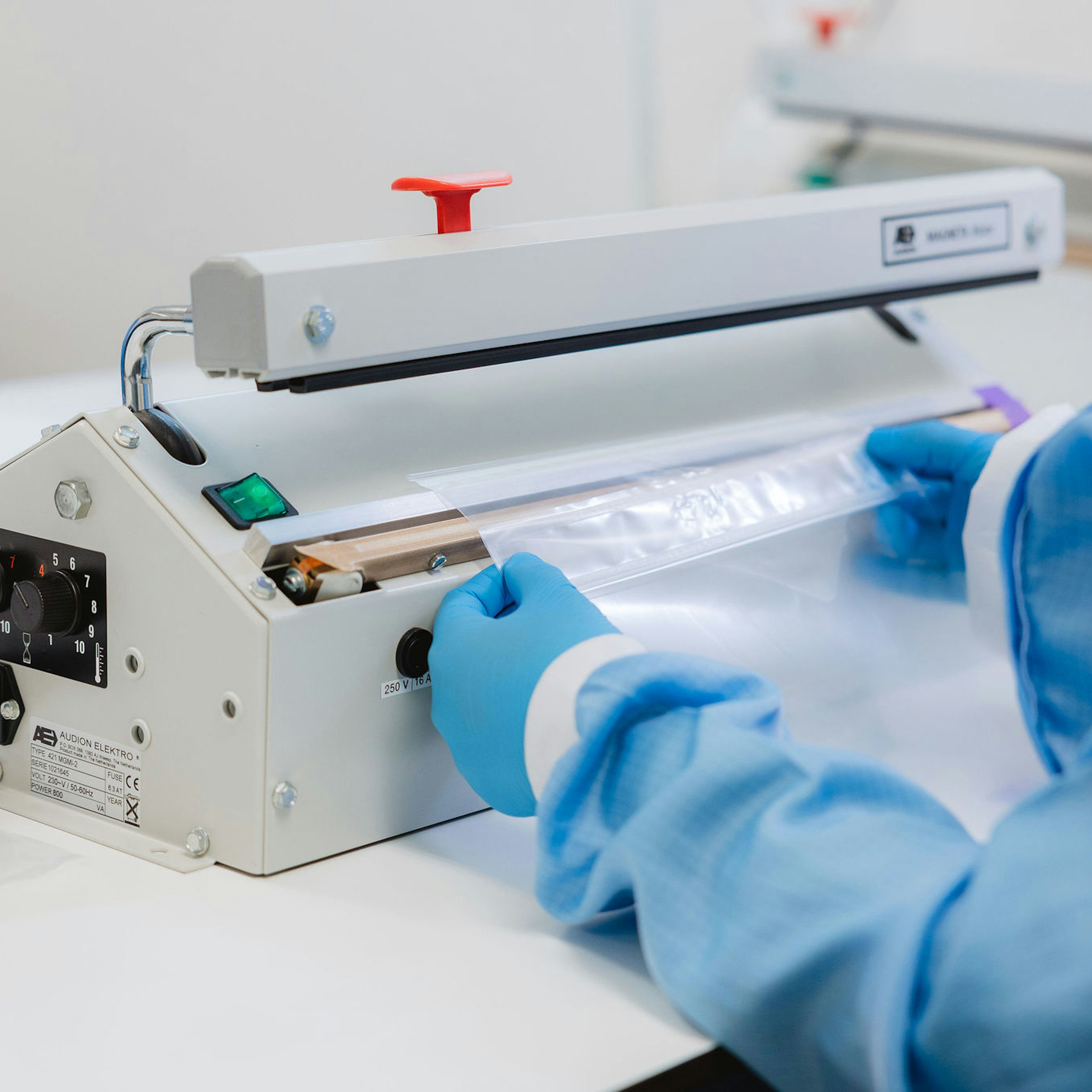 Een medewerker in een blauwe beschermende jas en nitrilhandschoenen sealt een transparant plastic zakje met een Audion Elektro impulssealer in een schone productieomgeving.