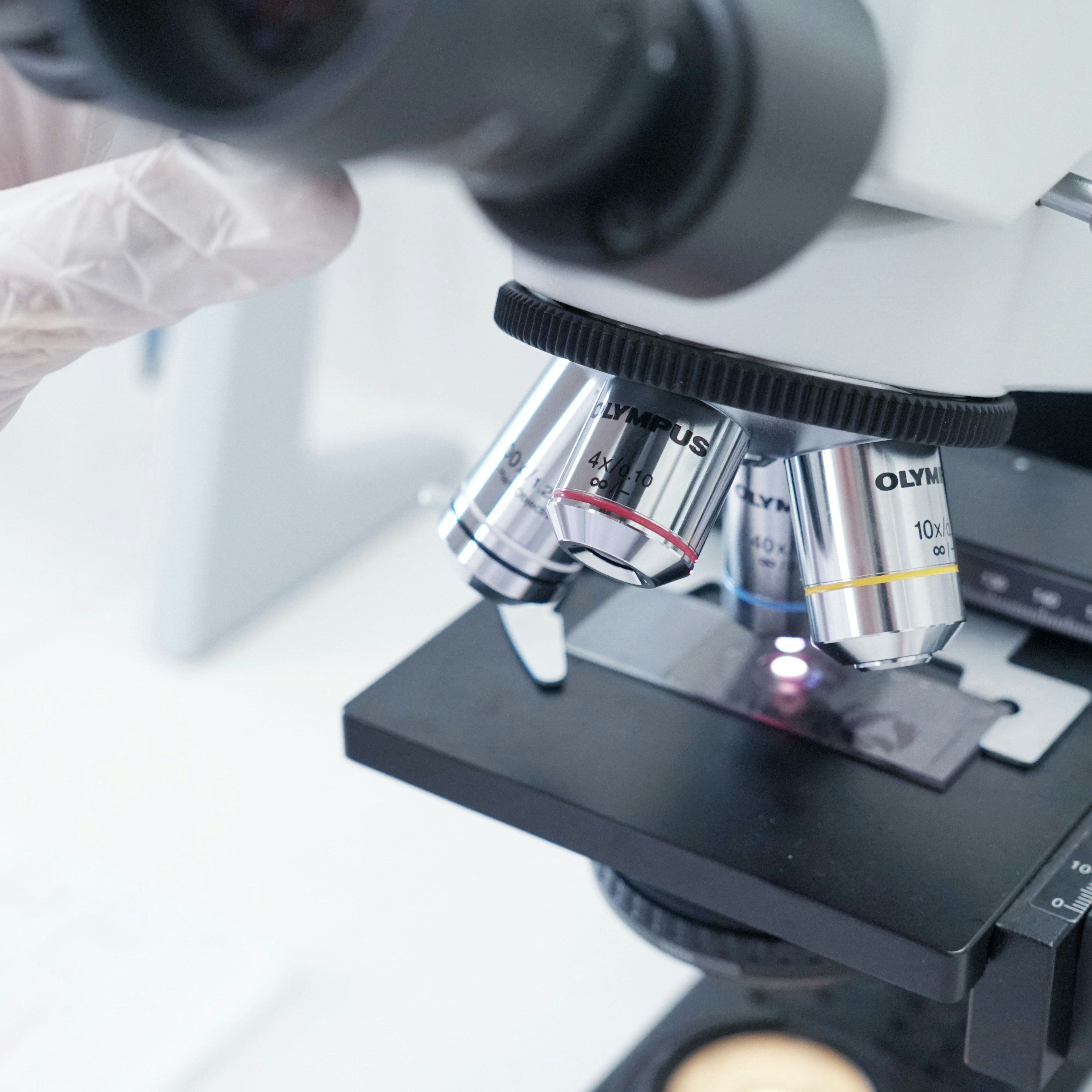 A close-up of a laboratory microscope, focused on its revolving nosepiece with several silver objective lenses of different magnifications (4x, 10x, 40x), marked with colored rings. A specimen slide sits on the black mechanical stage with its measurement scale visible. In the upper left, a gloved hand reaches toward the eyepieces. The bright white background keeps all attention on the precision instrument and its metallic details.