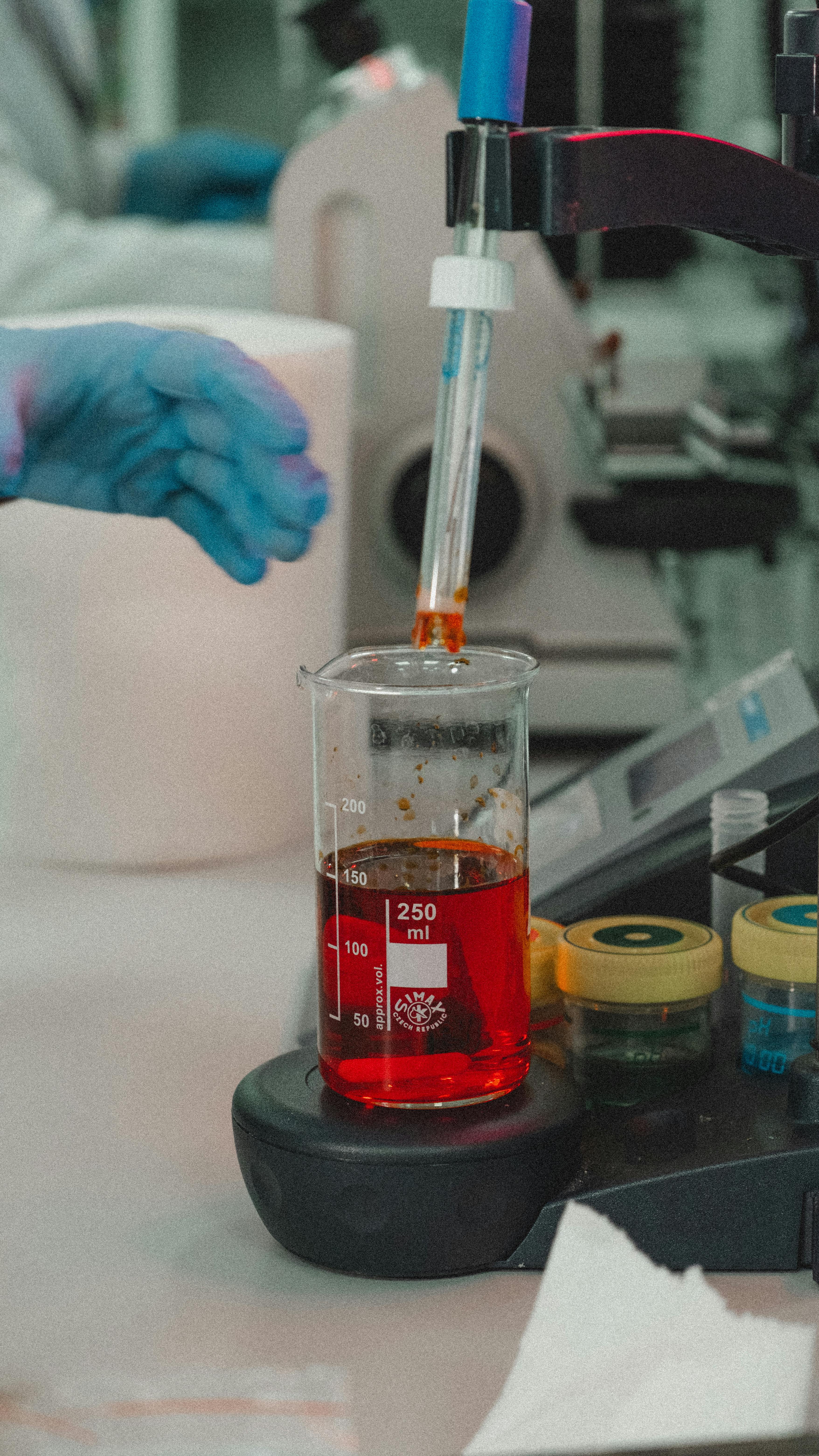 A close-up of a laboratory experiment centered on a 250 ml beaker containing a vivid red liquid, placed on what appears to be a black magnetic stirrer. A probe or electrode (possibly a pH meter) is submerged in the liquid, held by a stand. On the left, a blue-gloved hand reaches toward the beaker. On the right are two small jars with yellow caps containing dark samples. The background is blurred with lab equipment in cool tones, making the red liquid stand out strongly as the focal point of the composition.
