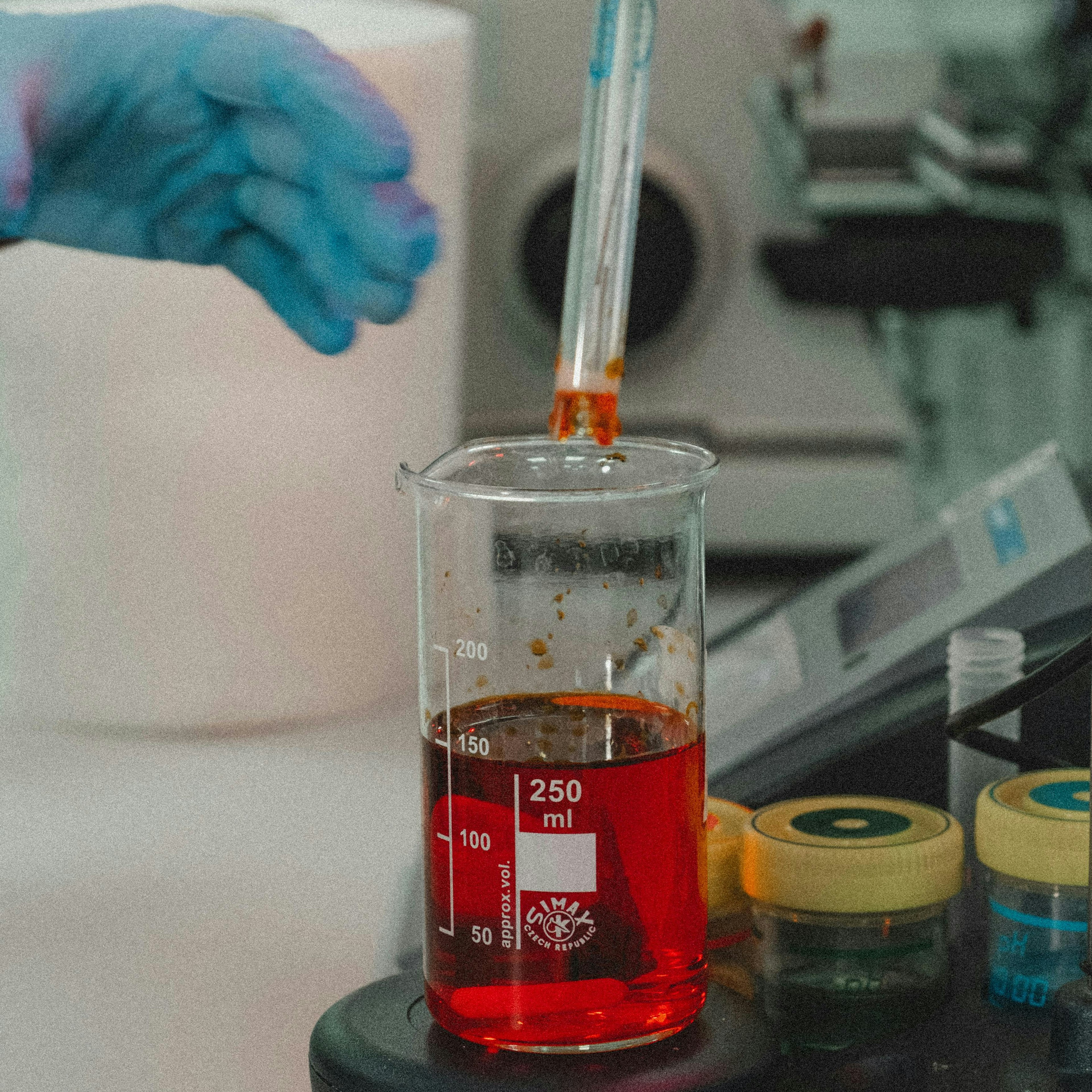 A close-up of a laboratory experiment centered on a 250 ml beaker containing a vivid red liquid, placed on what appears to be a black magnetic stirrer. A probe or electrode (possibly a pH meter) is submerged in the liquid, held by a stand. On the left, a blue-gloved hand reaches toward the beaker. On the right are two small jars with yellow caps containing dark samples. The background is blurred with lab equipment in cool tones, making the red liquid stand out strongly as the focal point of the composition.