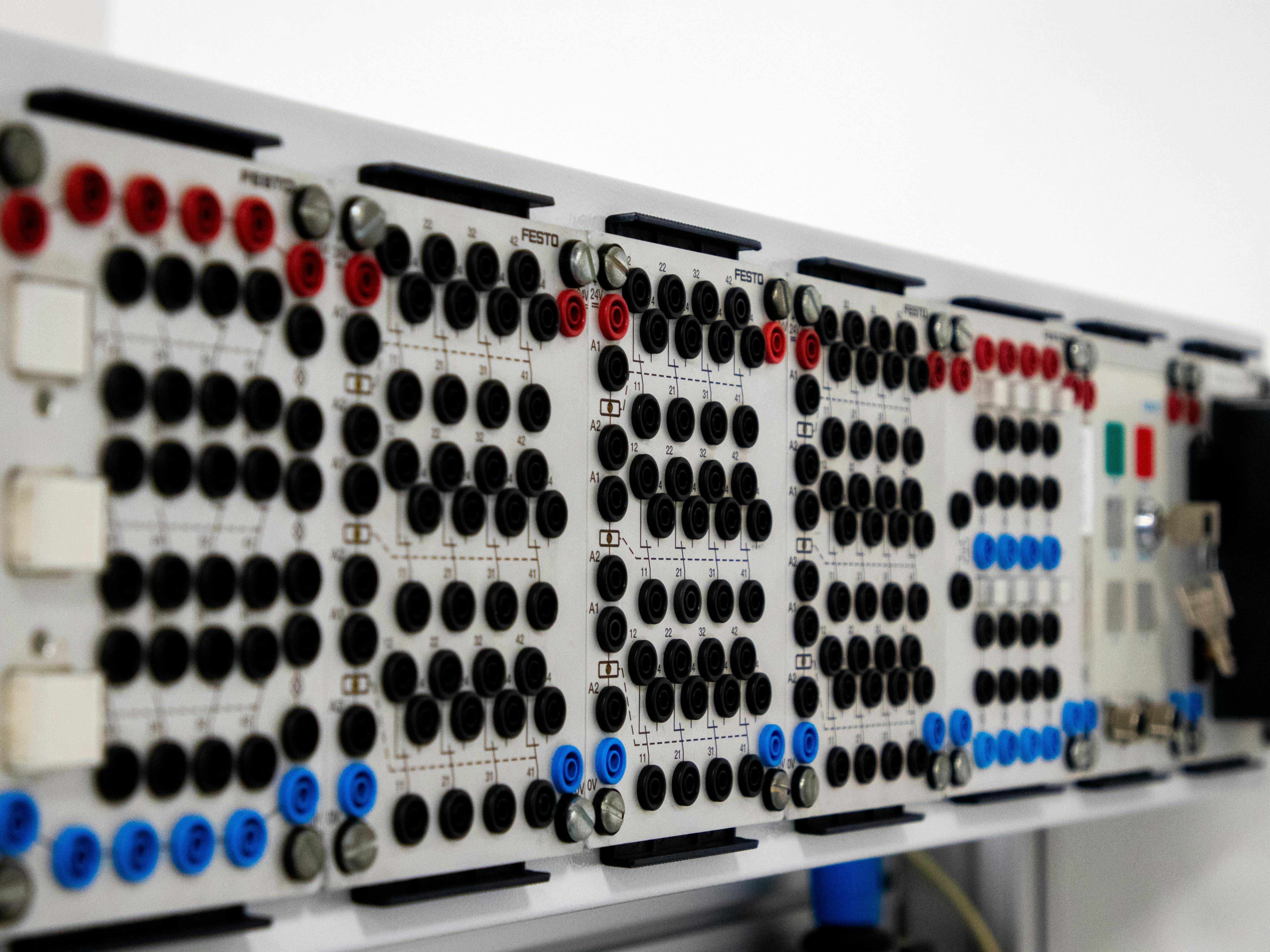 A close-up, angled view of an electrical training panel or relay control board, likely FESTO educational equipment used for teaching electrical engineering and automation. The board holds a row of white modules mounted on rails, each covered with arrays of black, red, and blue connection sockets (banana jacks) for wiring circuits. Faint circuit diagrams are printed on each module. On the right side there are colored push buttons (green and red) and what appear to be key switches. The shallow depth of field keeps the front modules sharp while the rest recede into a clean white background.