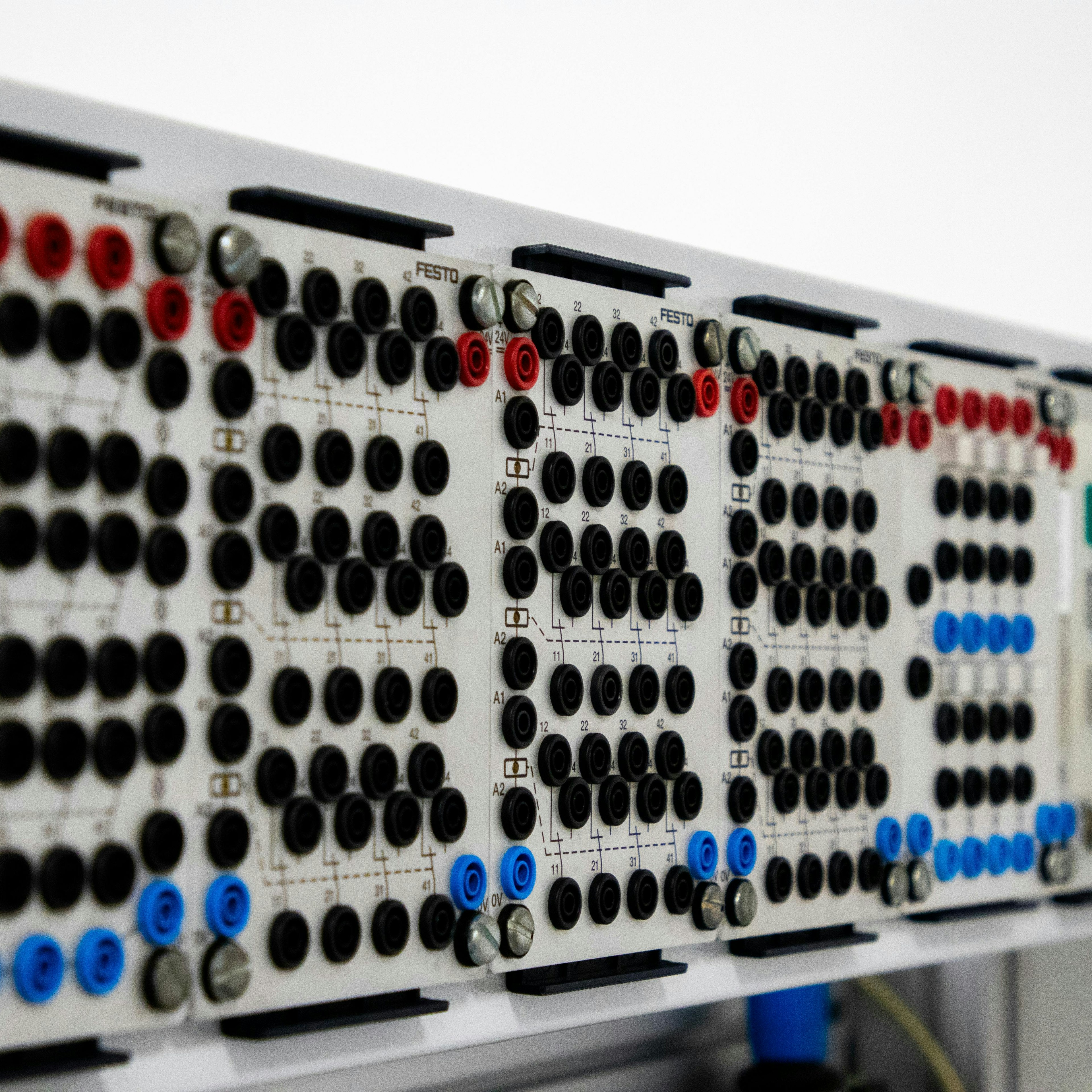 A close-up, angled view of an electrical training panel or relay control board, likely FESTO educational equipment used for teaching electrical engineering and automation. The board holds a row of white modules mounted on rails, each covered with arrays of black, red, and blue connection sockets (banana jacks) for wiring circuits. Faint circuit diagrams are printed on each module. On the right side there are colored push buttons (green and red) and what appear to be key switches. The shallow depth of field keeps the front modules sharp while the rest recede into a clean white background.