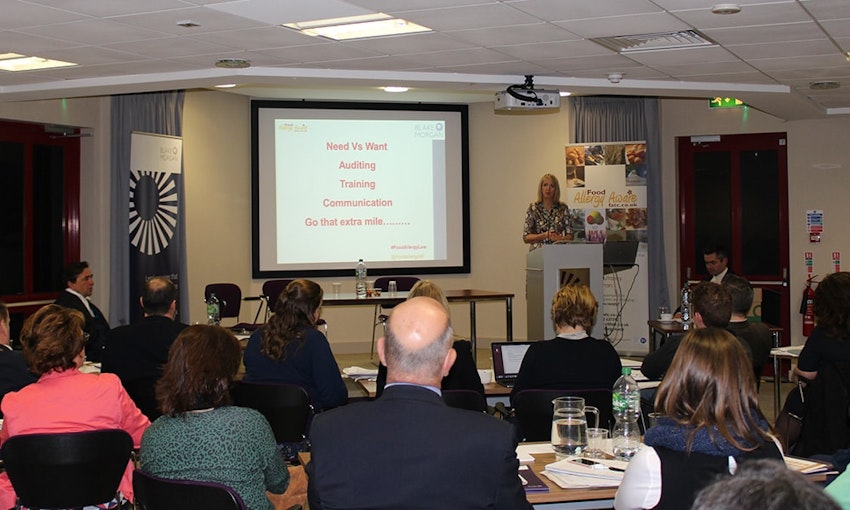 Caroline Benjamin presenting before the mock trial - Image Copyright Food Allergy Aware Caroline Benjamin presenting to audience projector screen has red writing which says needs vs wants auditing training communication go that extra mile