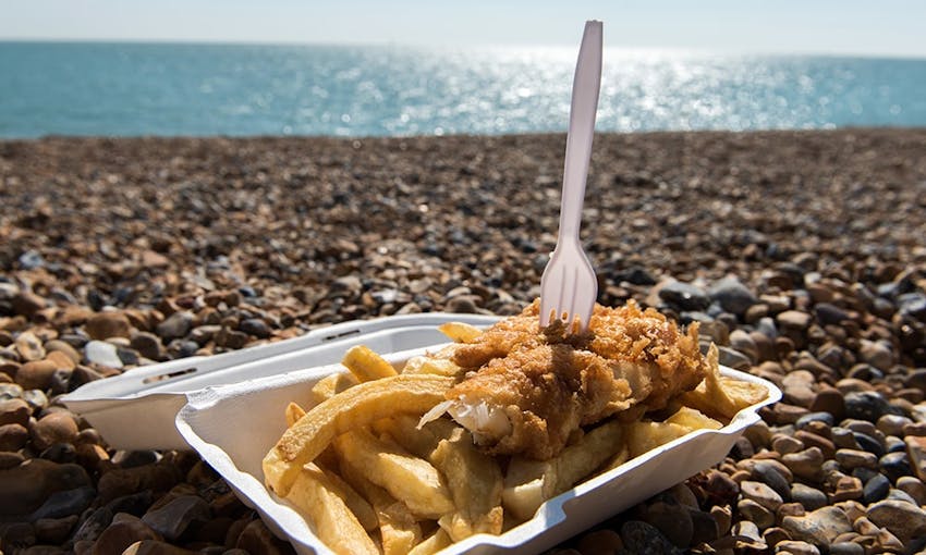 a cardboard tray of traditional british fish and chips with a plastic fork in the middle of a half eaten fish on a shingled beach infront of glistening blue sea 