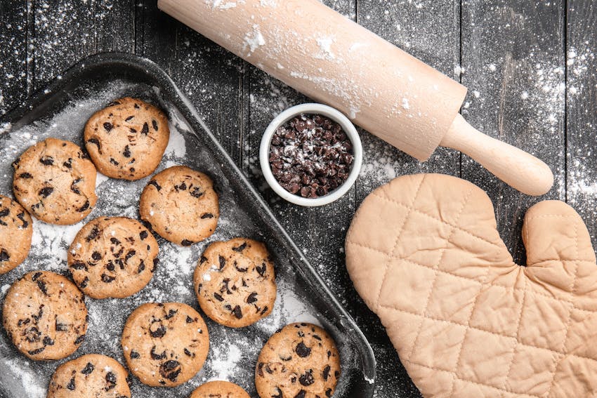 a tray of 12 chocolate chip cookies next to a small dish of chocolate chips, a rolling pin and an oven glove all dusted with flour 