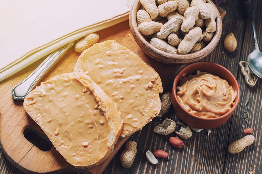 two slices of peanut butter toast on a wooden chopping board next to a dish of peanuts and a dish of crunchy peanut butter