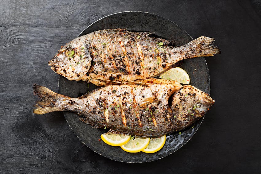 two grilled fishes garnished in herbs and spices on a bed of sliced lemon in a black bowl 