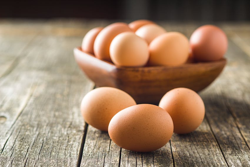 three eggs on a wooden table in front of a square wooden bowl filled with eggs