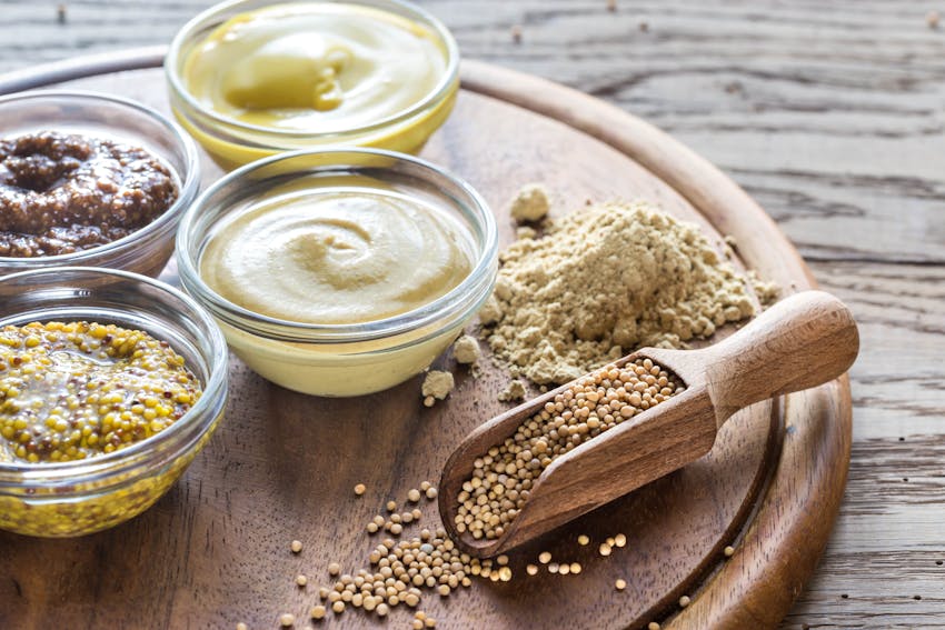 various glass dishes of mustard on a circular wooden chopping board with mustard seeds on a wooden spoon 