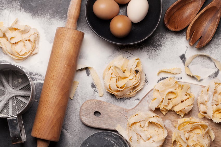 a rolling pin and chopping board placed next to fresh tagliatelle and four eggs covered with flour   