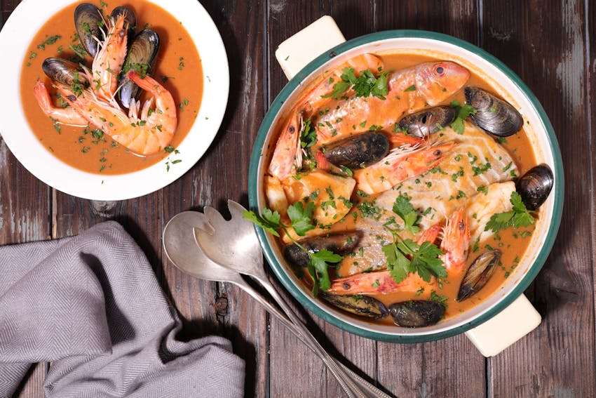 a large hotpot of bouillabaisse next to a smaller serving dish of bouillabaisse topped with fresh herbs 
