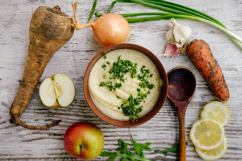 A bowl of apple and parsnip soup topped with herbs surrounded by fresh from the ground root vegetables
