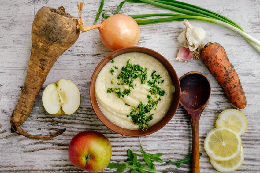 A bowl of apple and parsnip soup topped with herbs surrounded by fresh from the ground root vegetables