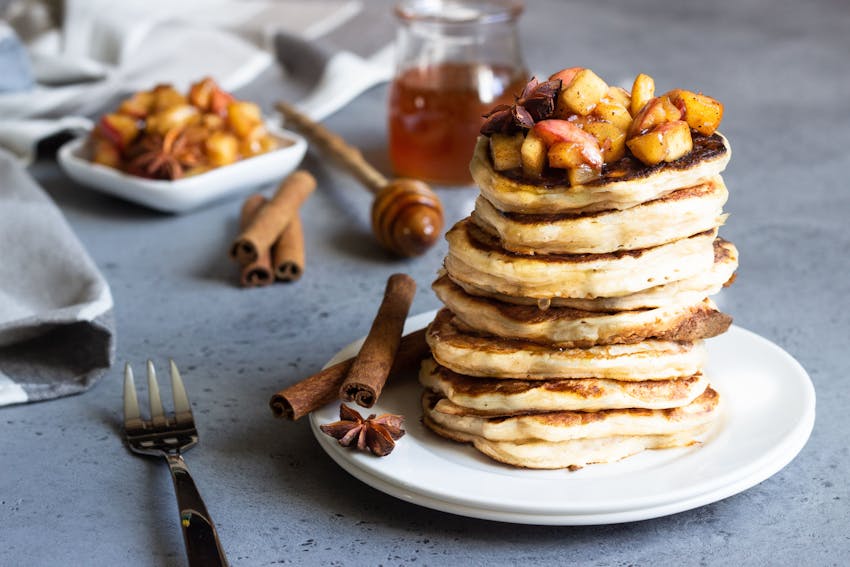 a white plate with a stack of eight pancakes with stewed apples on top and presented with two cinnamon sticks