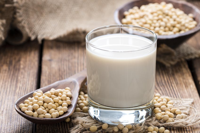 small glass of soya milk next to a wooden spoon of soya beans on a dark wooden table with decorative cloth 