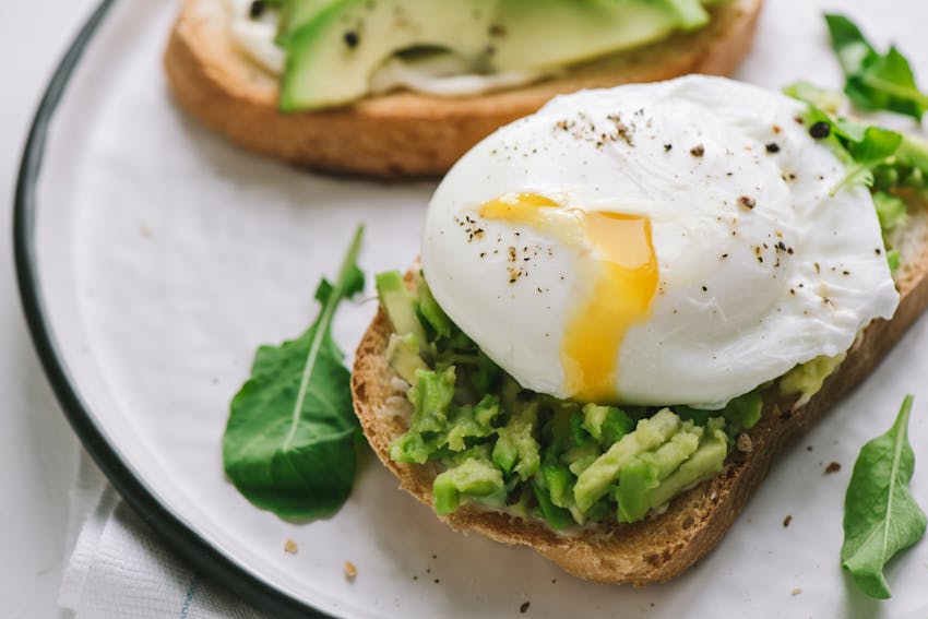 two slices of brown bread topped with smashed avocado, a poached egg with yolk oozing out with pepper and spinach on a white plate 