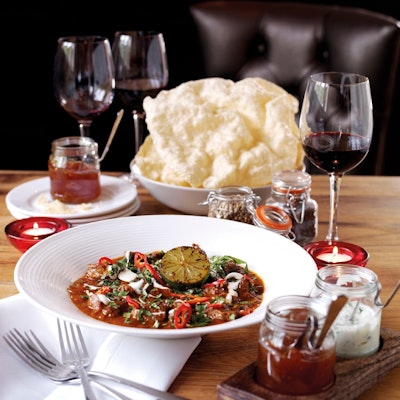 dish of welsh lamb curry served on a table with two small glass jars of condiments a bowl of poppadoms and three glasses or red wine 