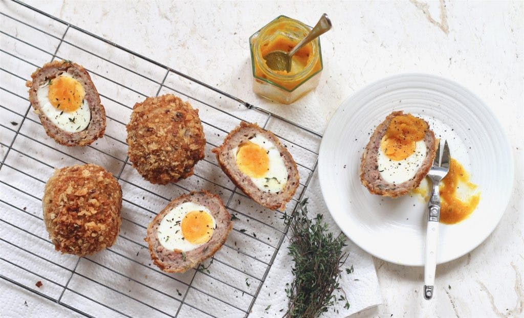 one scotch egg halved on a white saucer with a dollop of mustard and two scotch eggs halved on a wire cooling rack dusted with black pepper and fresh herbs 