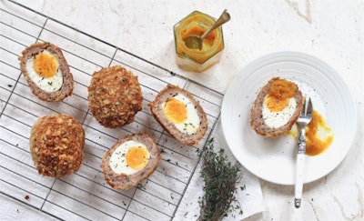 one scotch egg halved on a white saucer with a dollop of mustard and two scotch eggs halved on a wire cooling rack dusted with black pepper and fresh herbs 