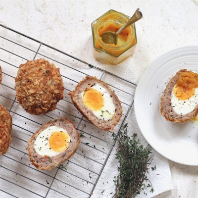 one scotch egg halved on a white saucer with a dollop of mustard and two scotch eggs halved on a wire cooling rack dusted with black pepper and fresh herbs 