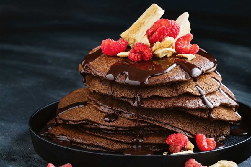 a stack of seven gluten free chocolate pancakes drizzled with chocolate sauce and raspberries 