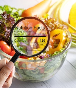 hand holding a magnifying glass illuminates nutrition and calorie information infront of a bowl of fresh crispy salad next to a calculator 