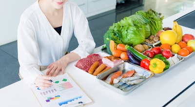 woman in a white shirt sits at a white table filled with a range of different fruits as she overlooks food information and charts 