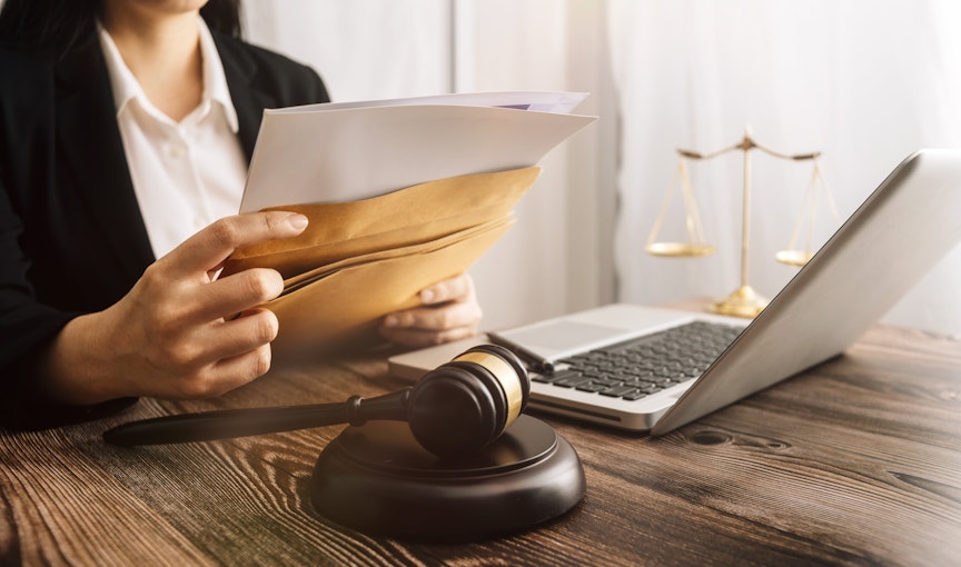 lawyer sits at a desk with laptop and dark wooden law gavel as she slips documents into a brown envelope 